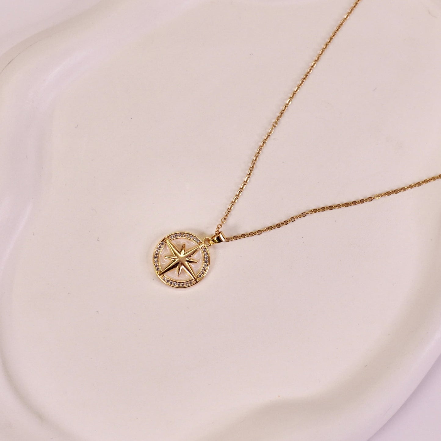 A gold-colored pendant necklace with a compass design on a luminous pearl, displayed on a white plate.