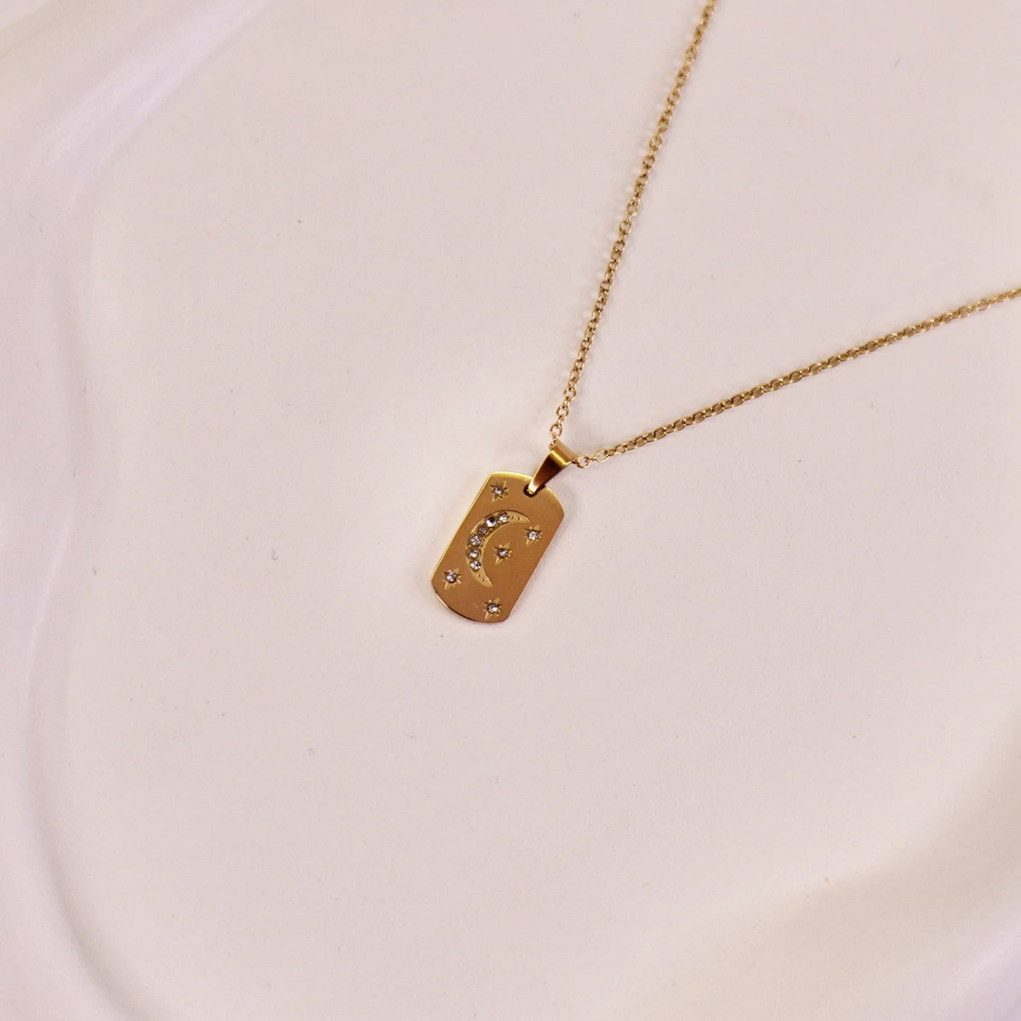 A gold-colored pendant necklace with a crescent moon design on a rectangular plaque, featuring micro-studded accents, displayed on a white plate.