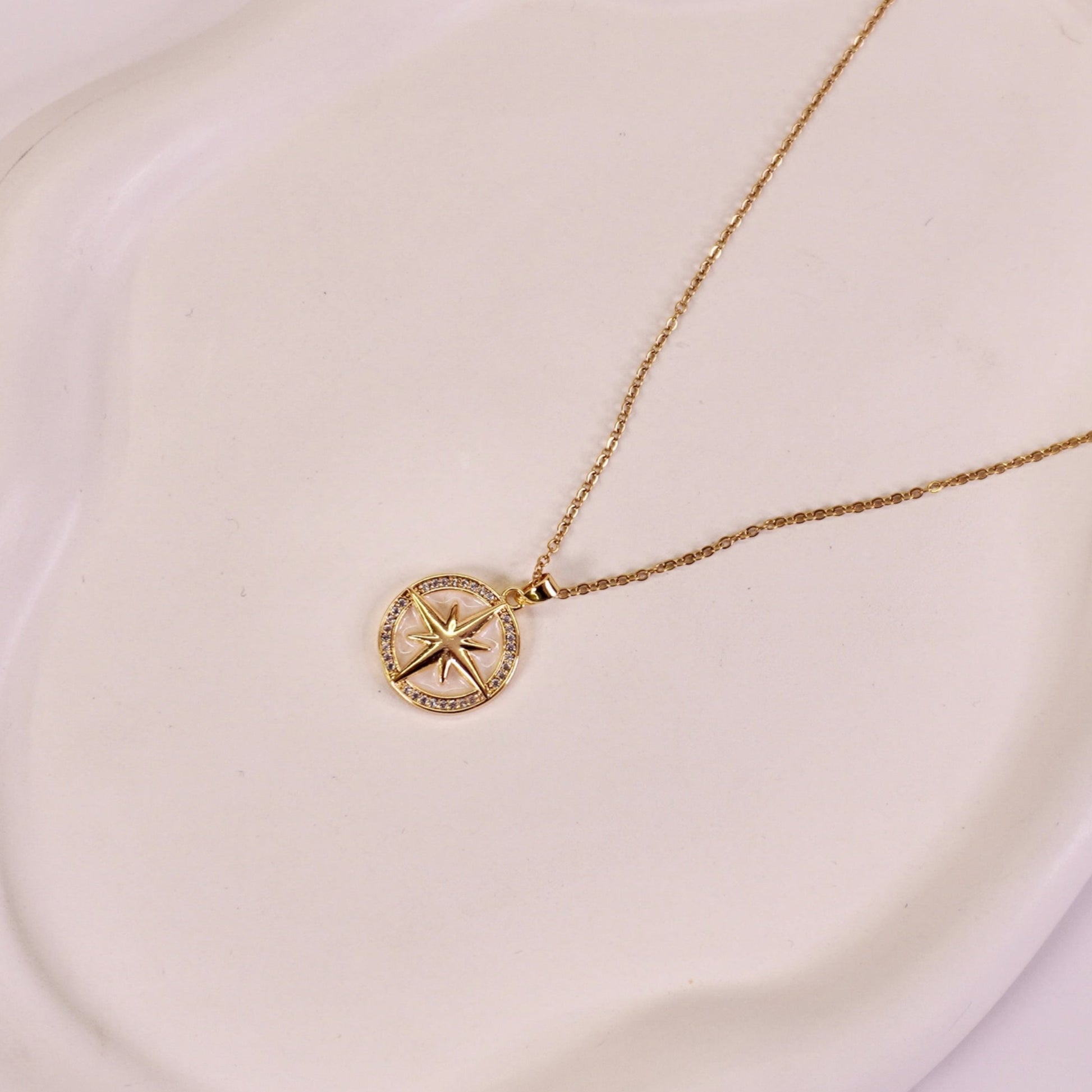 A gold-colored pendant necklace with a compass design on a luminous pearl, displayed on a white plate.