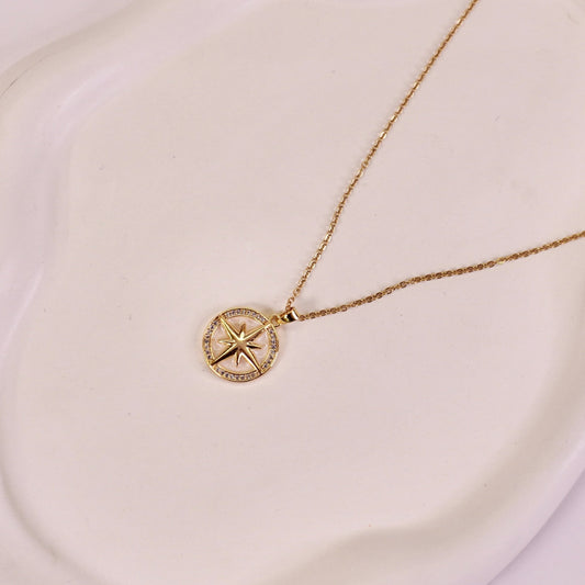 A gold-colored pendant necklace with a compass design on a luminous pearl, displayed on a white plate.