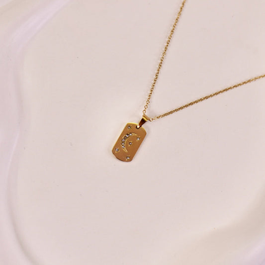 A gold-colored pendant necklace with a crescent moon design on a rectangular plaque, featuring micro-studded accents, displayed on a white plate.